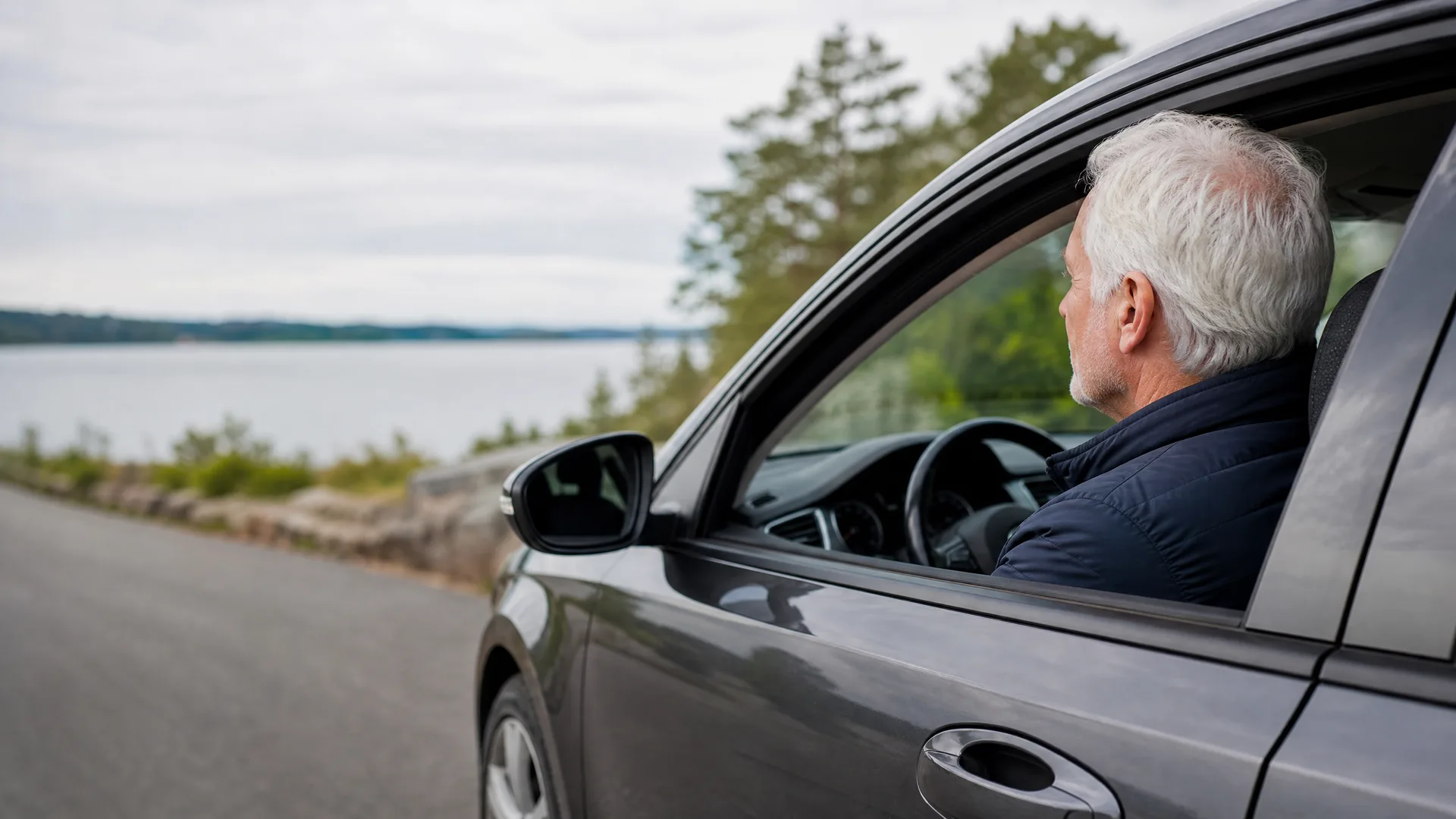 A gray-haired man sits in the driver's seat of a dark gray car parked beside a road, looking out the open window toward a body of water and trees.
