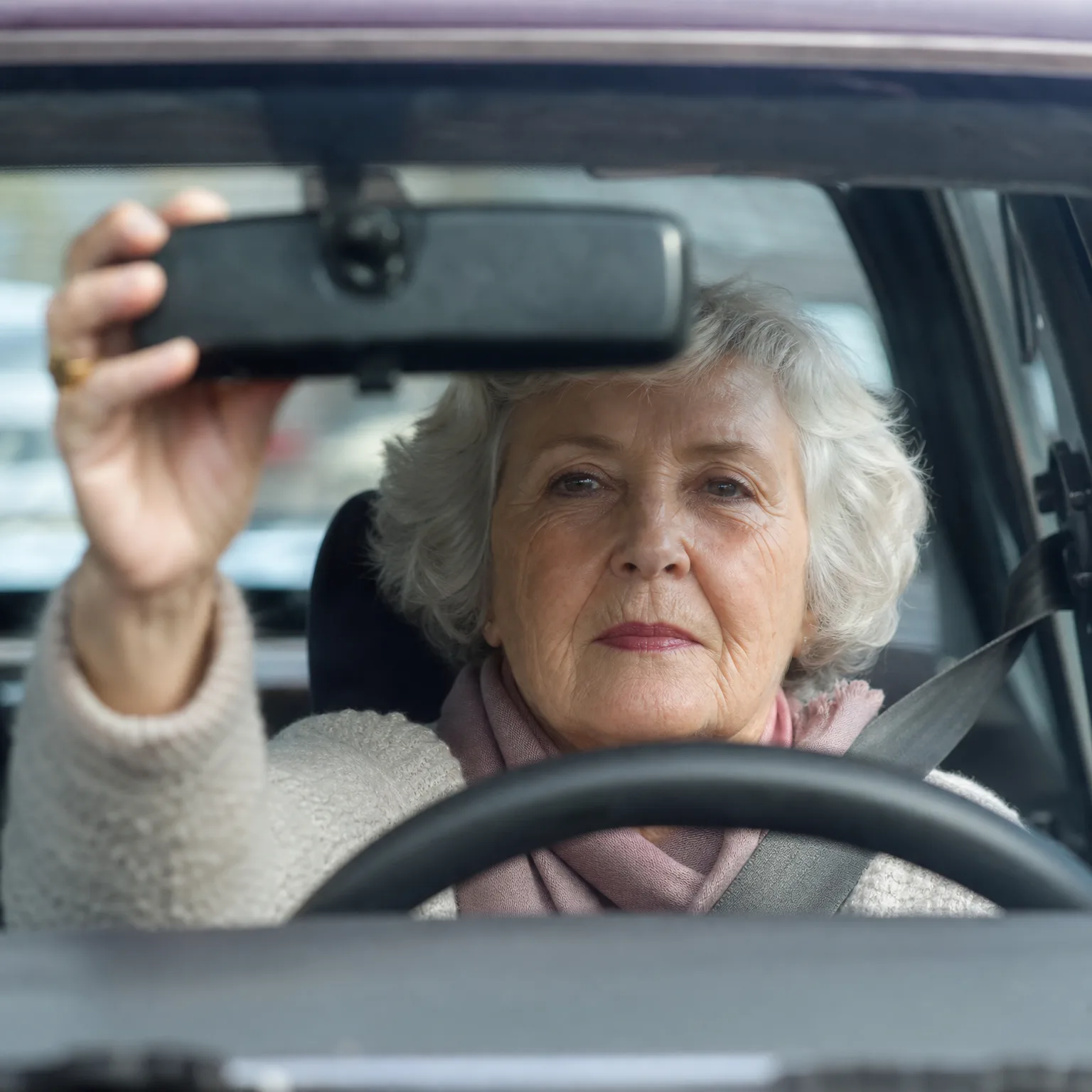An older woman with white hair and a pink scarf sits in the driver's seat of a car, reaching up to adjust the rearview mirror, seen through the windshield.